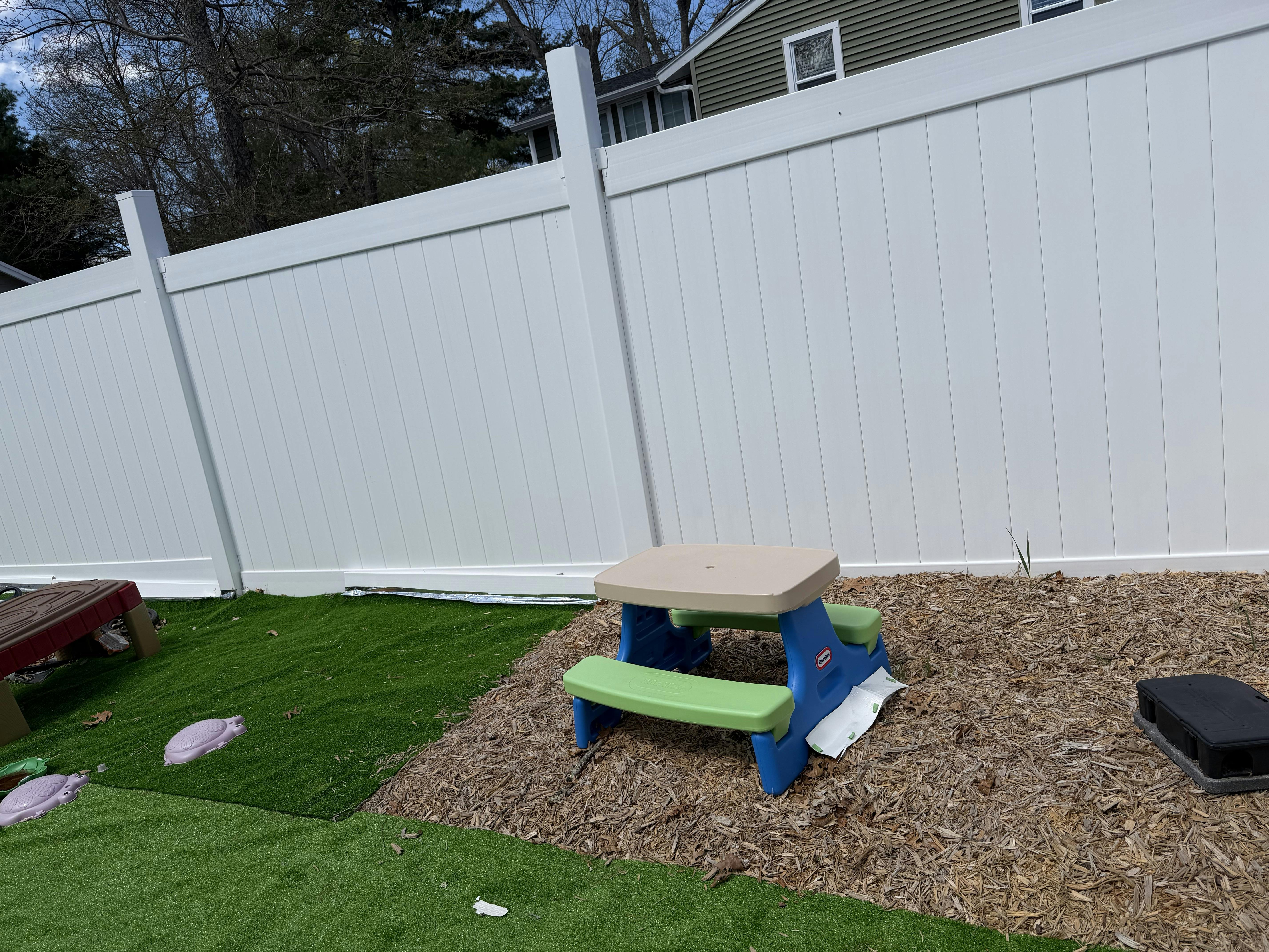 Child-sized picnic table on wood chips next to artificial turf and a white fence