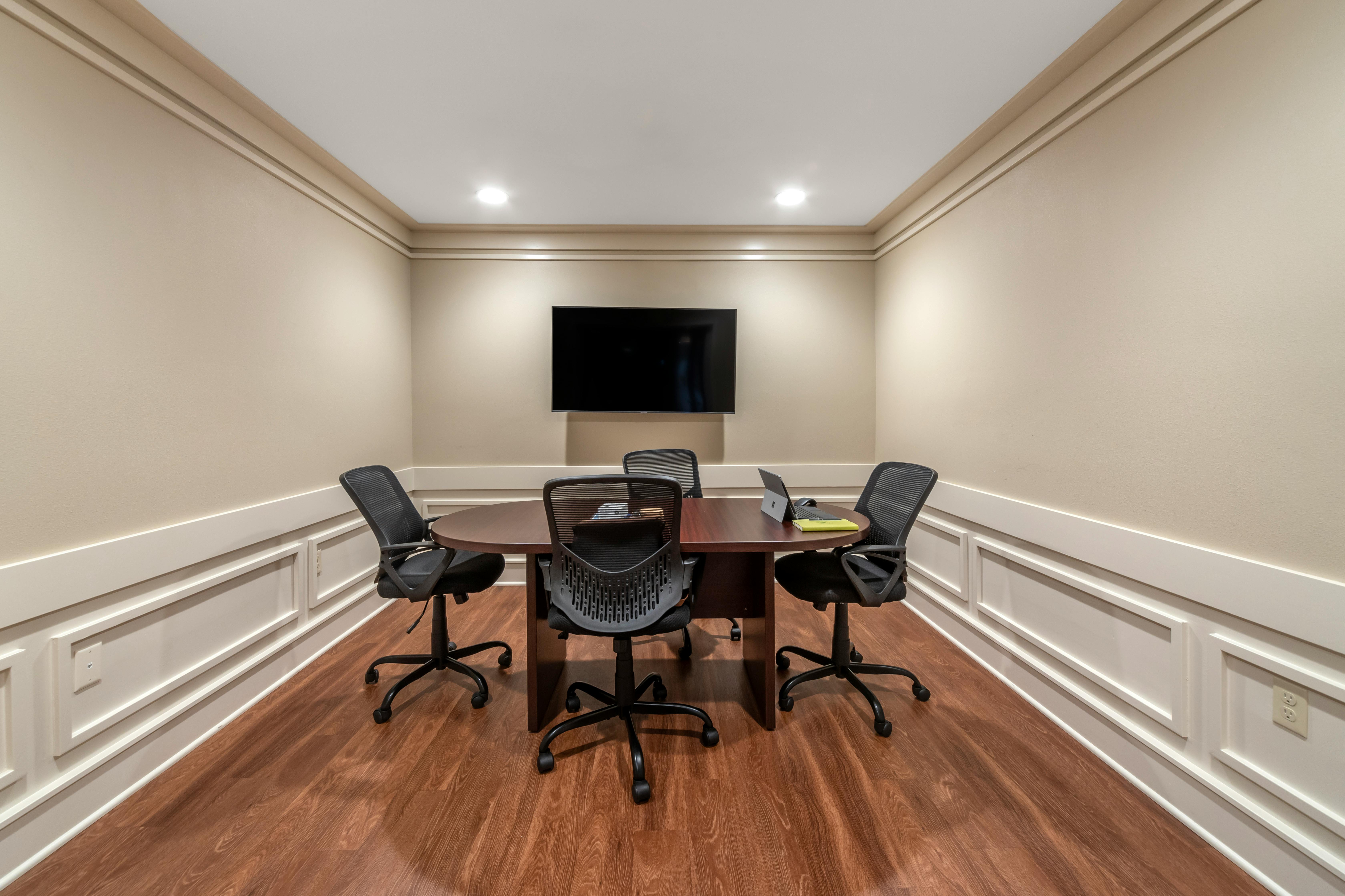 Small conference room with a round wooden table, four office chairs, and a wall-mounted TV