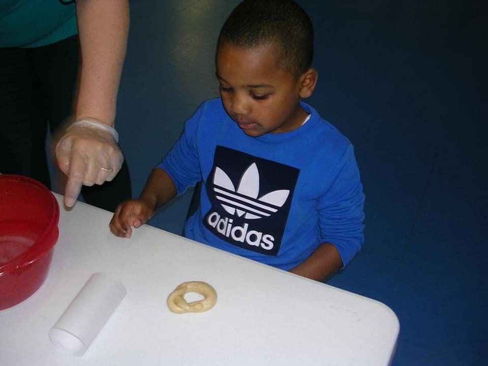 Child at a table with an adult's hand pointing to a small dough ring