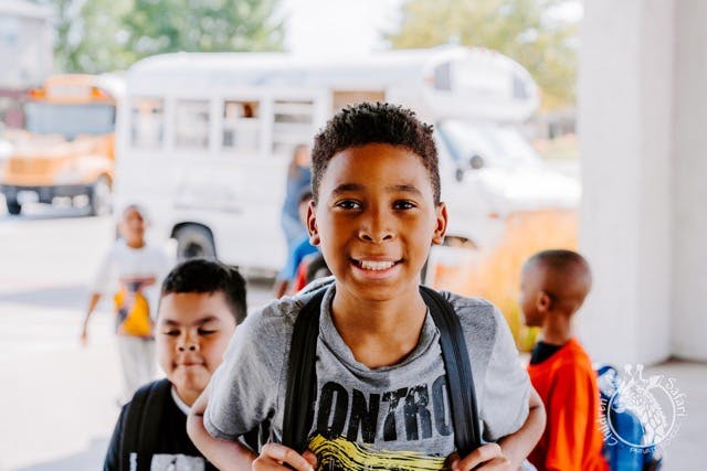 Child with backpack smiling at camera, other children and buses in background