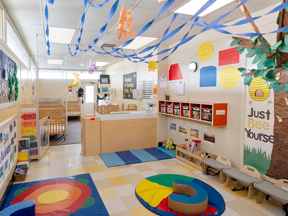 Classroom with cribs, playmats, and decorations including a tree cutout tree with "Just Bee Yourself"