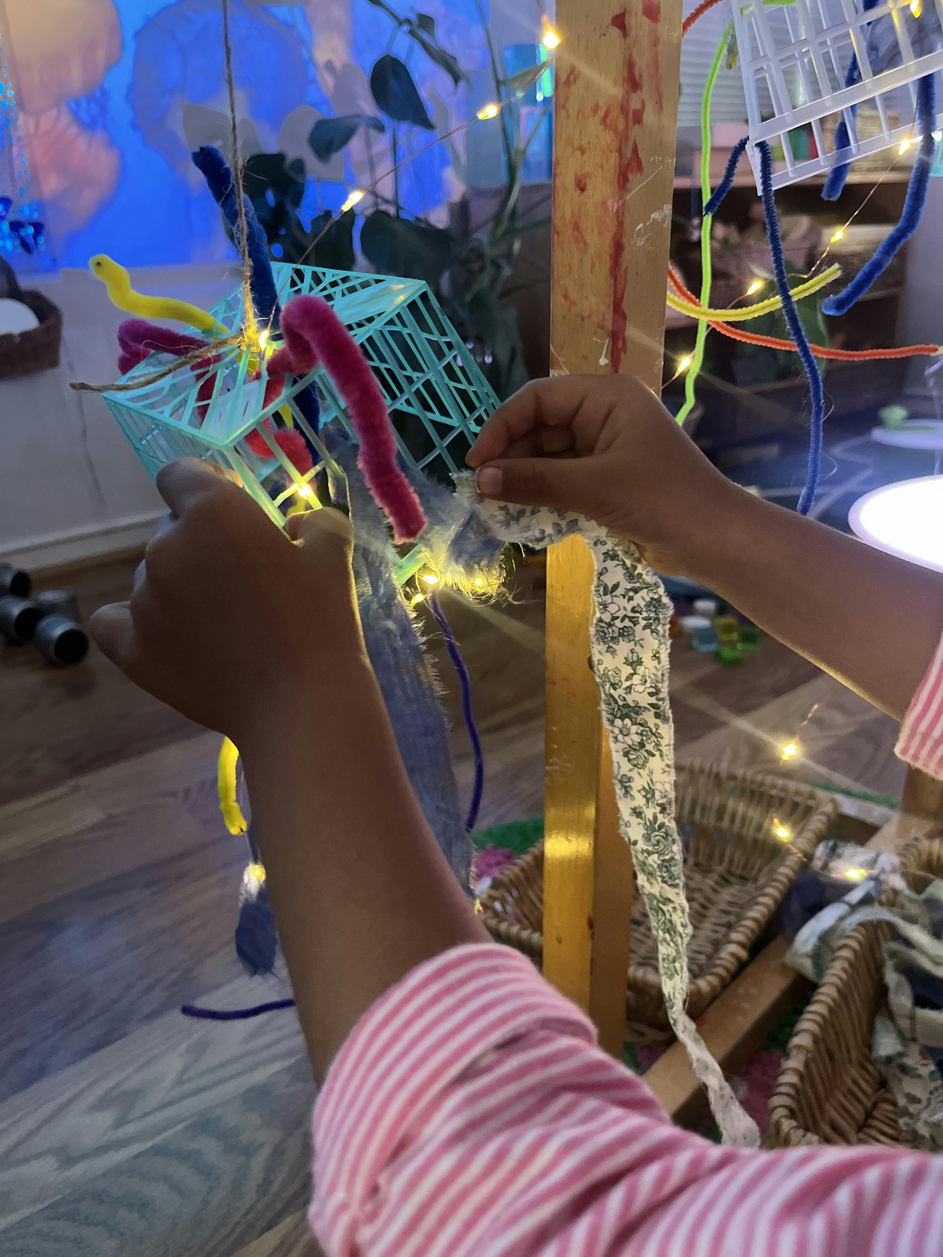 Child threading a ribbon through a plastic crate decorated with pipe cleaners