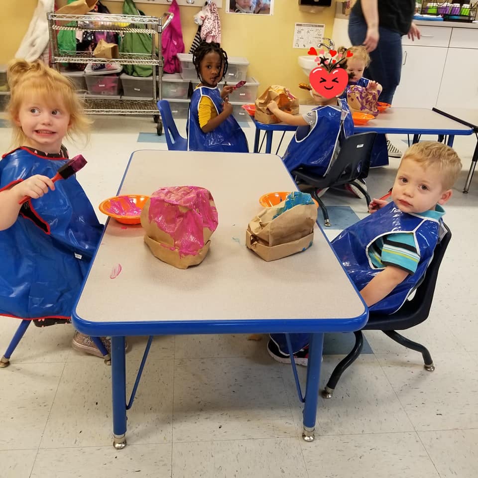 Children in blue smocks painting at tables in a classroom