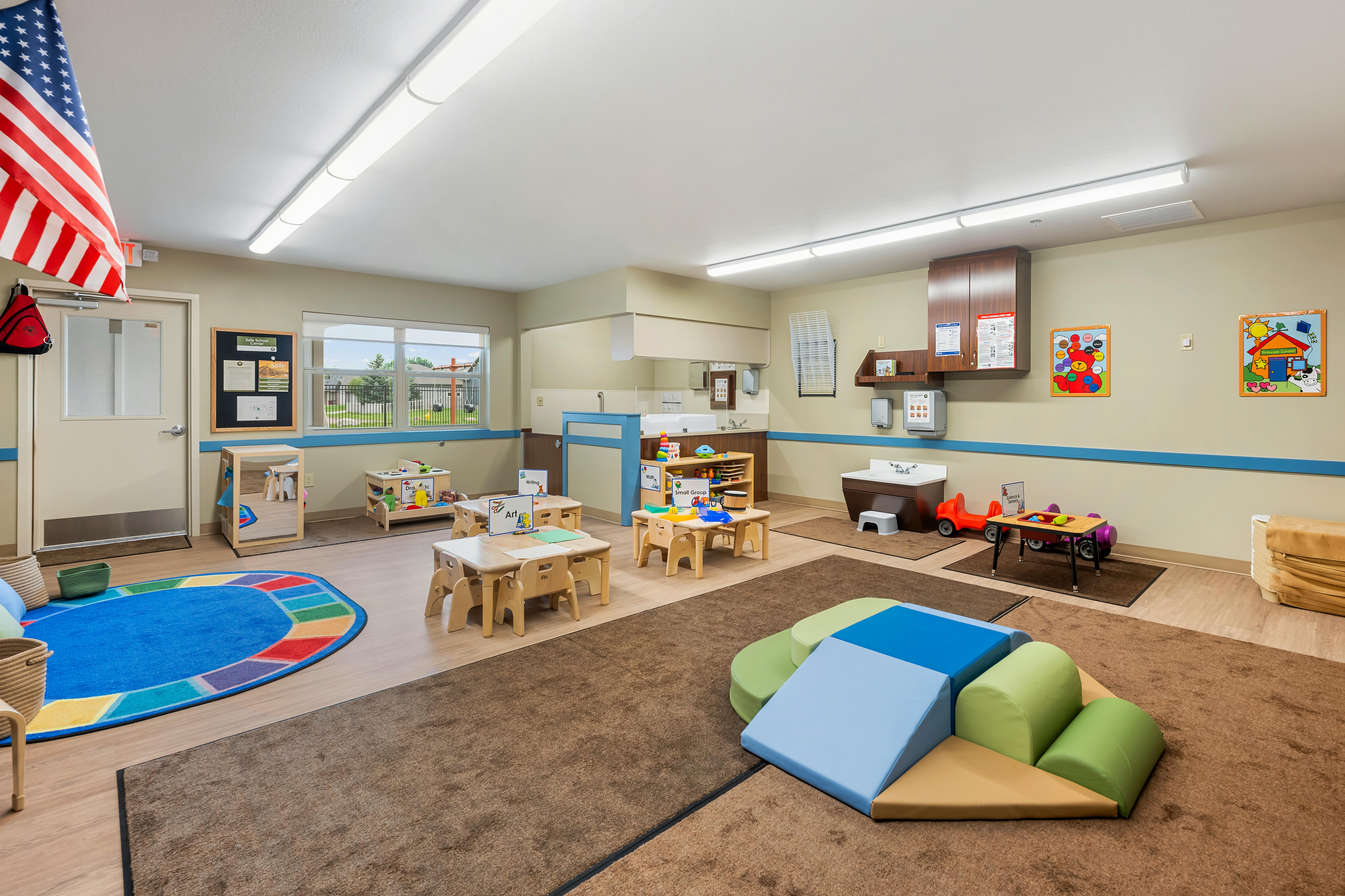 An empty early childhood classroom with small tables and chairs, a play area, and activity centers
