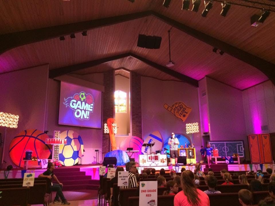 Stage with "Game On!" screen, large sports balls, and people in an audience