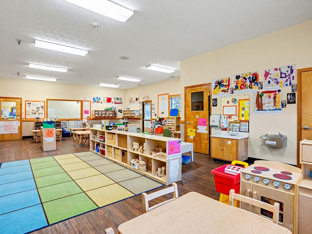 Classroom with colorful rug, small tables, chairs, and shelves with toys