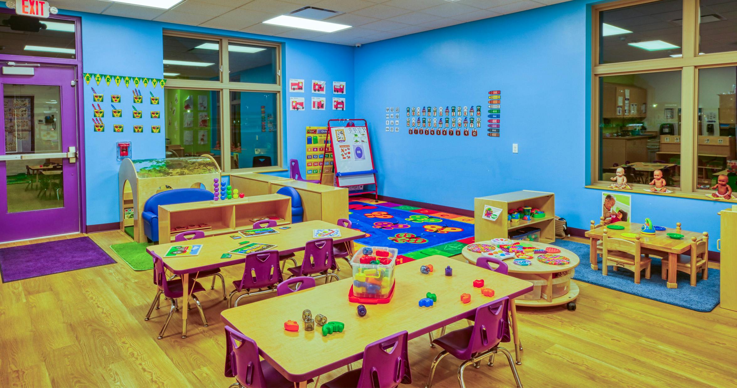 Colorful classroom with small tables and chairs, toys, and educational posters on blue walls