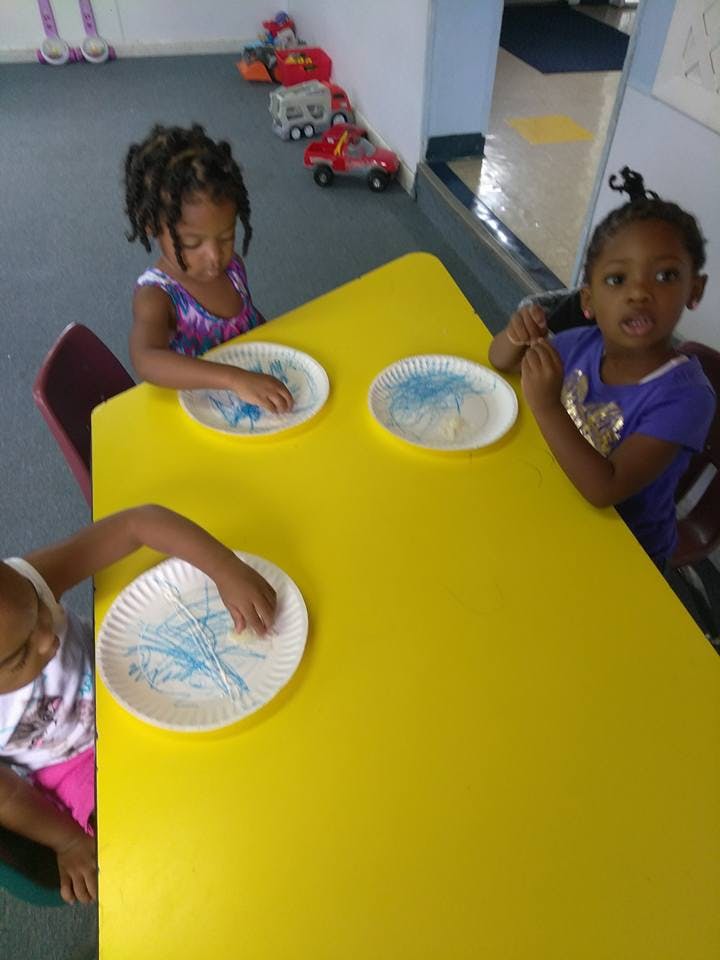 Three children at a yellow table with paper plates, one child looking up