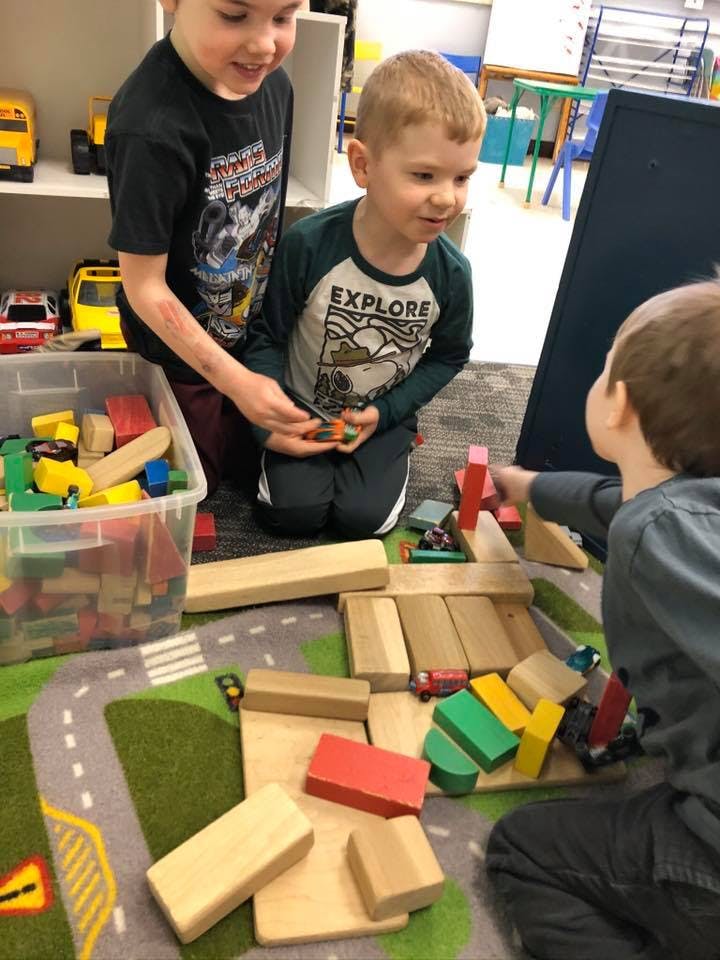 Three children playing with wooden blocks and toy cars on a rug