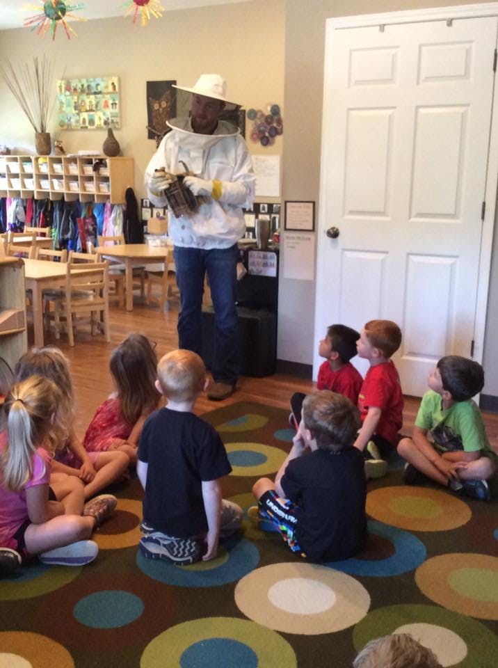 Person in beekeeper suit holding equipment, speaking to children sitting on floor