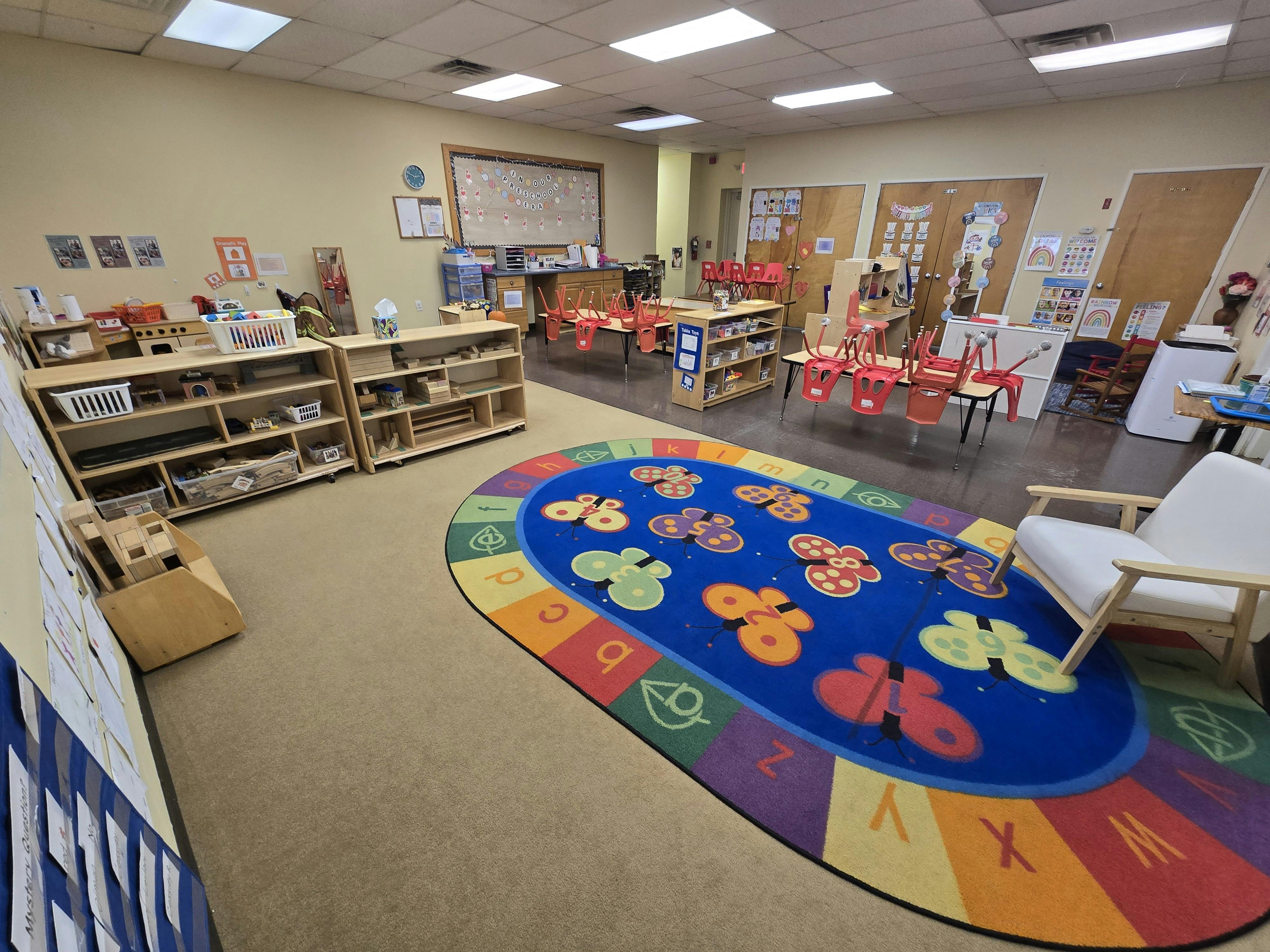 Classroom with small tables and chairs, shelves of toys, and a butterfly rug