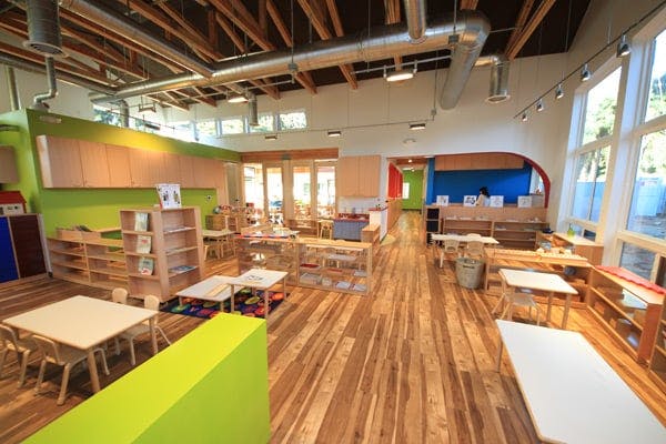 Empty classroom with small tables and chairs, wooden shelves, and large windows