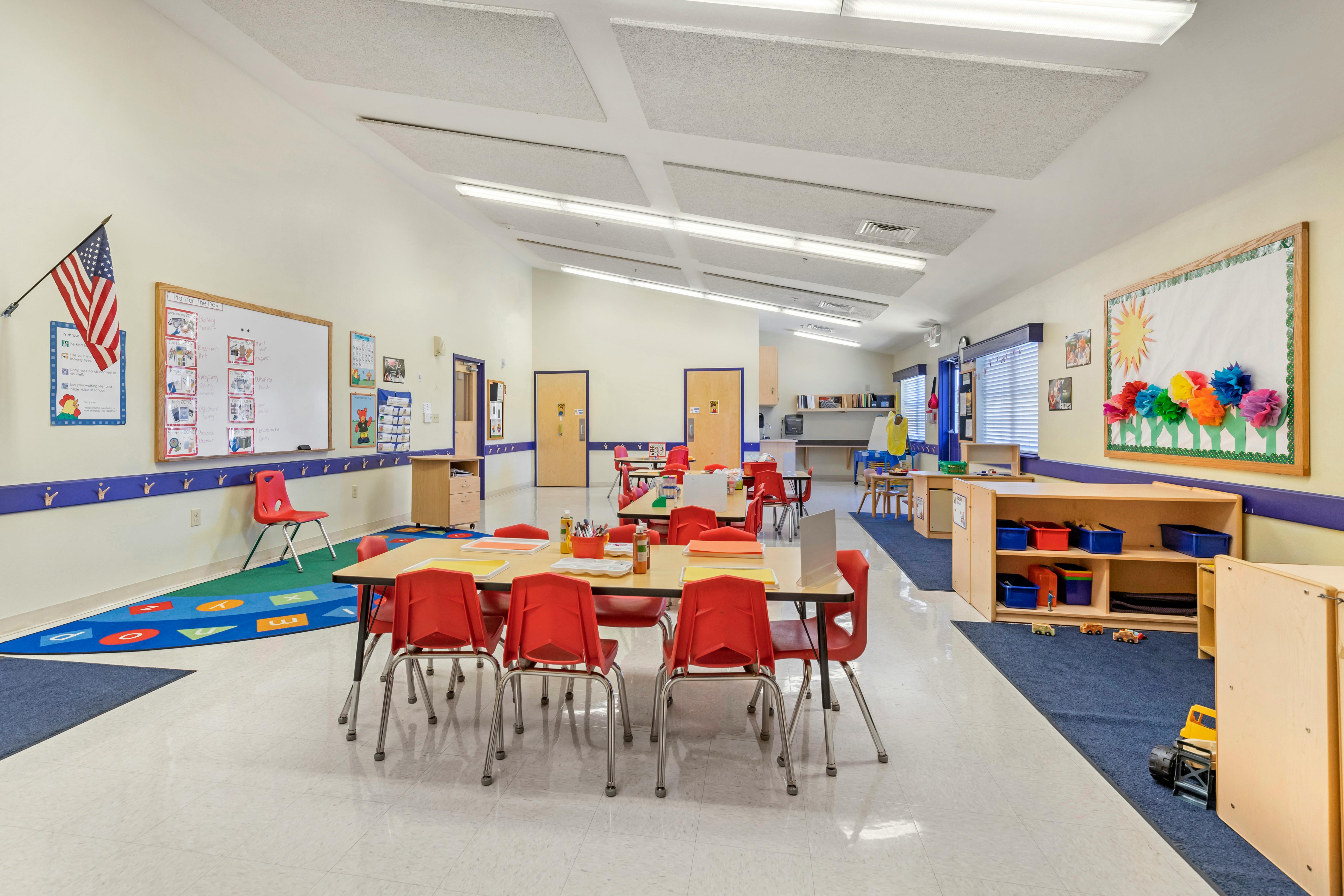 Classroom with small tables, red chairs, and a whiteboard with "Plan for the Day"