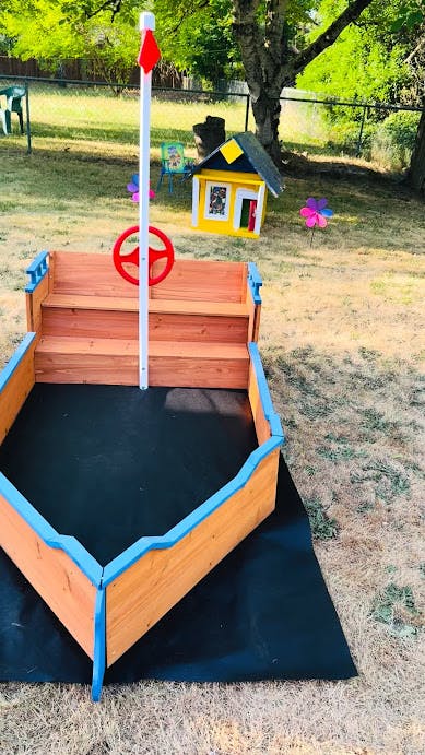 Wooden boat-shaped sandbox with a steering wheel and flag in a yard