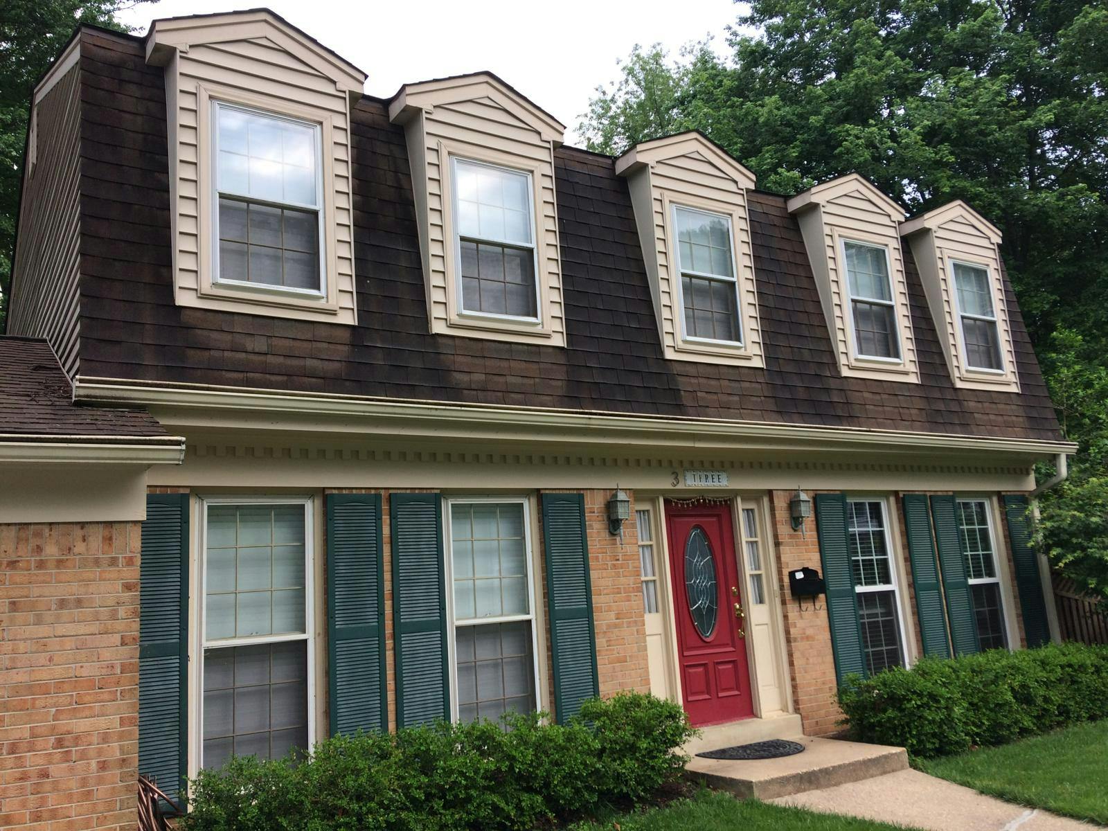 Brick house with brown shingle roof, three dormer windows, green shutters, red door