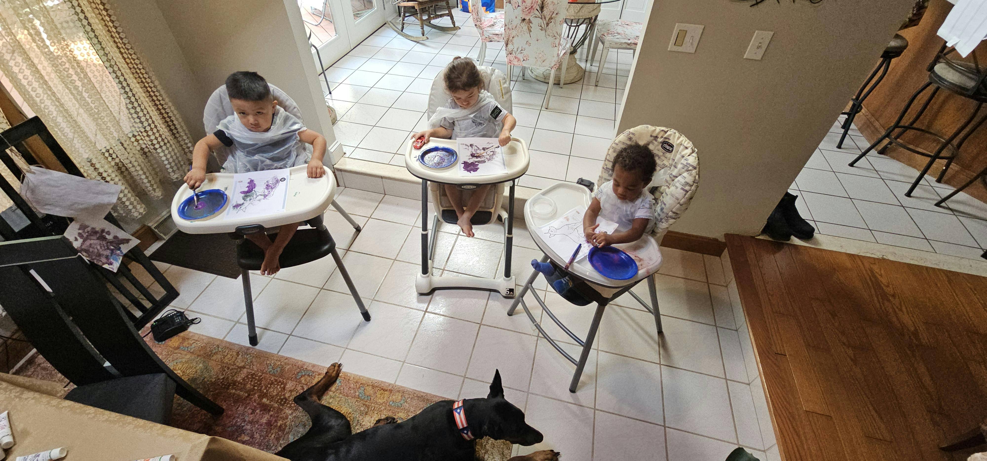 Three children in high chairs with a dog on the floor