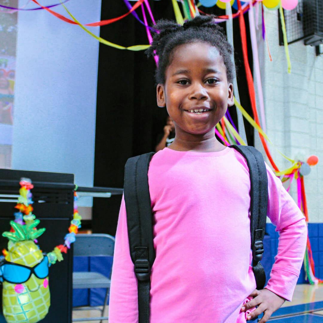 Child in a pink shirt wearing a backpack, smiling