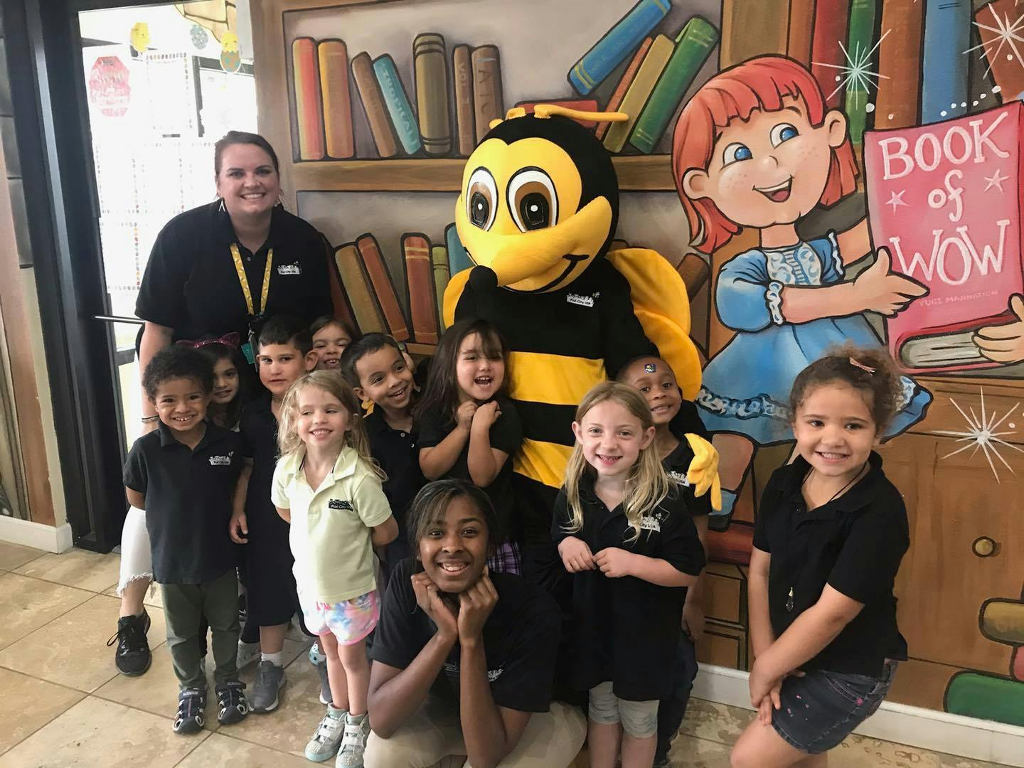 Adults and children posing with a bee mascot in front of a book-themed mural