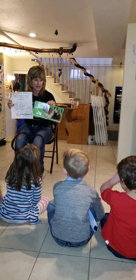 Person sitting on a chair holding a book and paper, three children sitting on floor