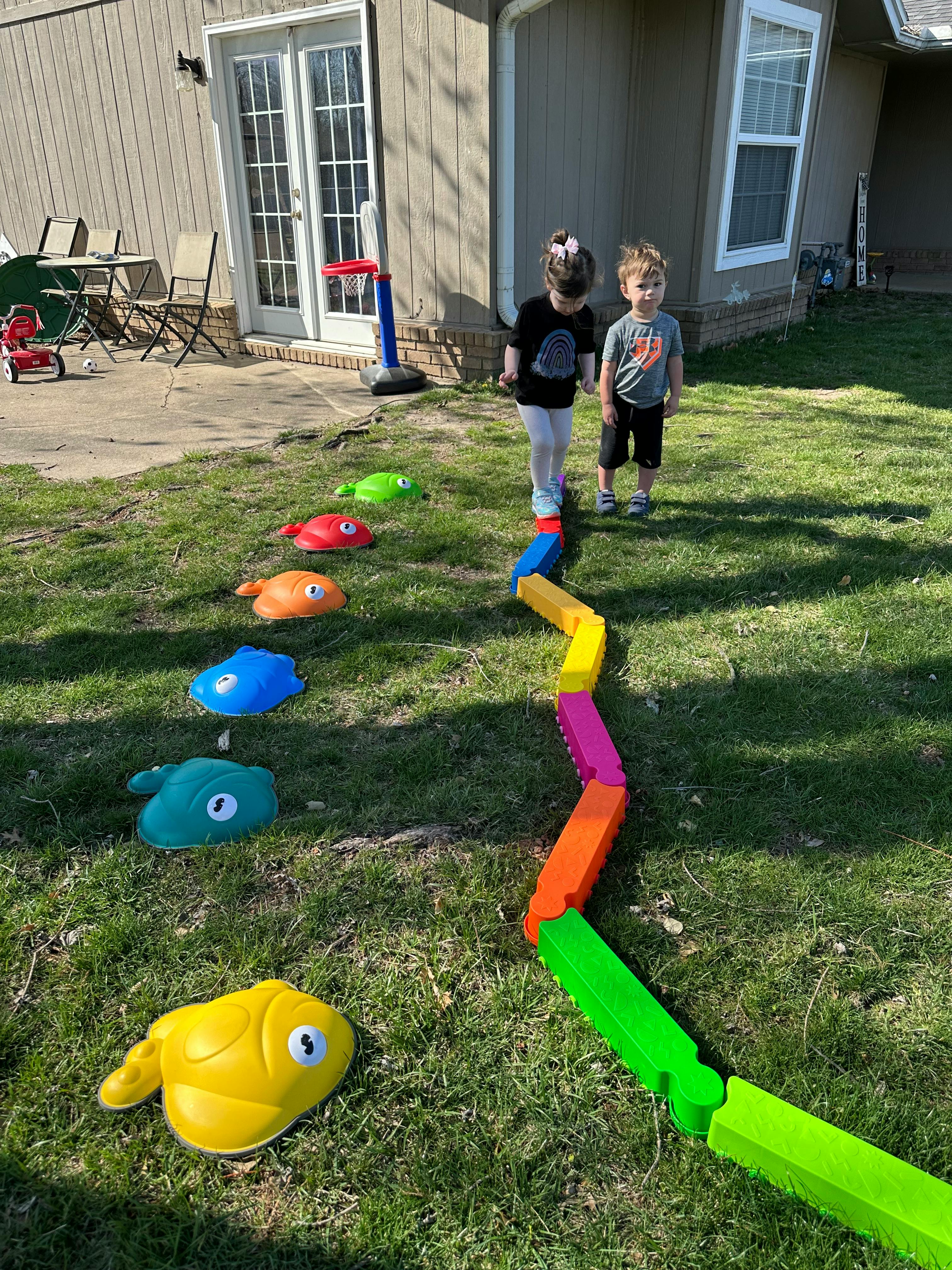 Two children walking on a colorful balance beam in a grassy yard