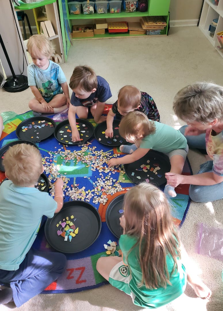 Group of children and an adult sorting colorful mosaic tiles into black trays on a rug