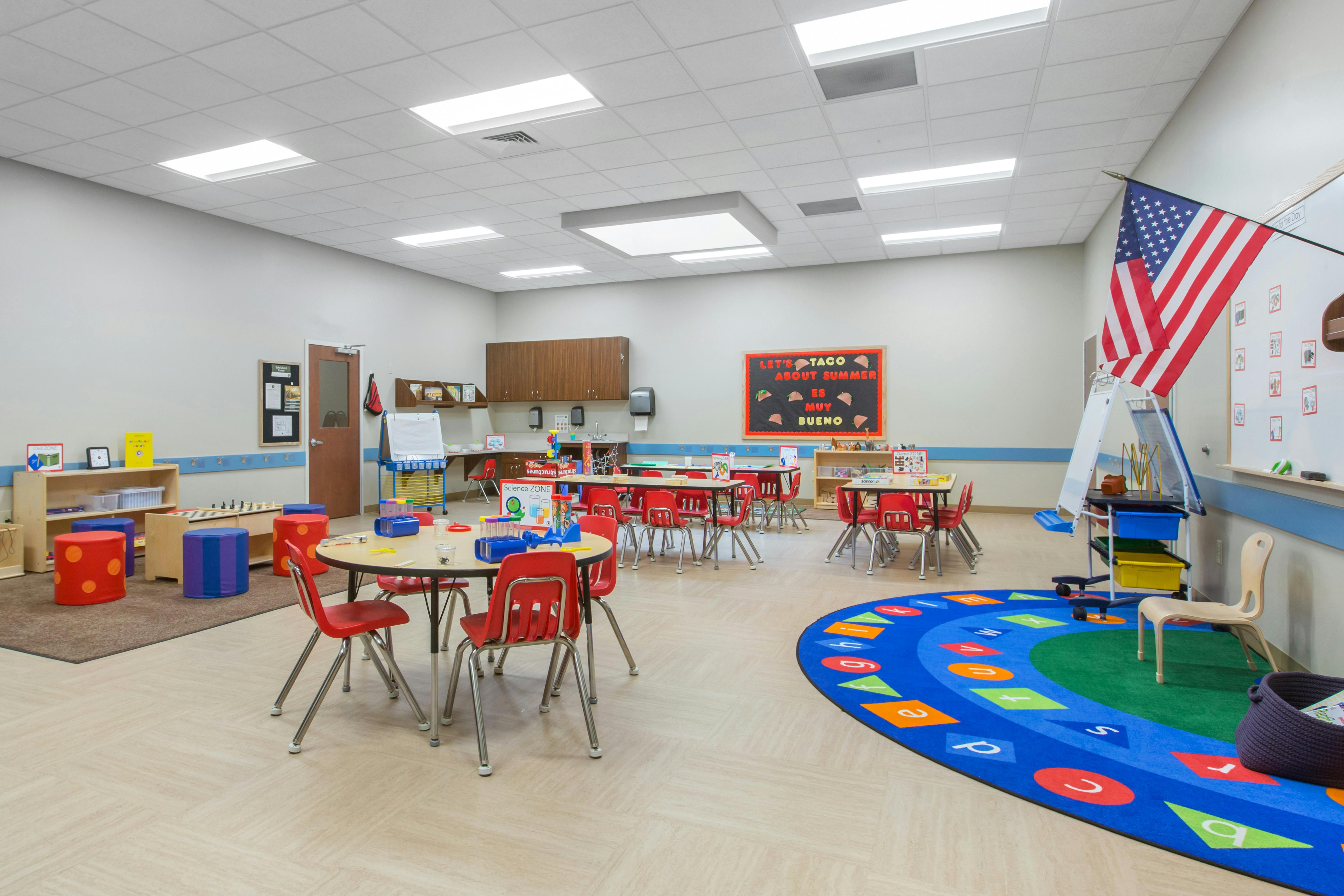 Classroom with small tables and chairs, a play rug with letters, and an American flag