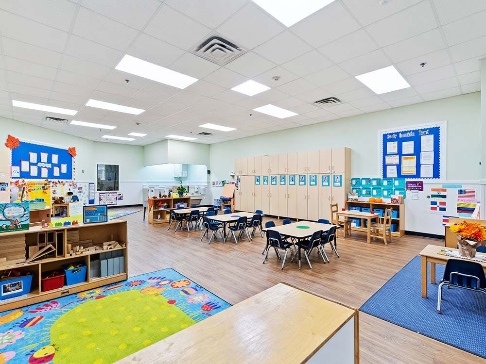 Empty classroom with small tables and chairs, shelves with toys, and bulletin boards