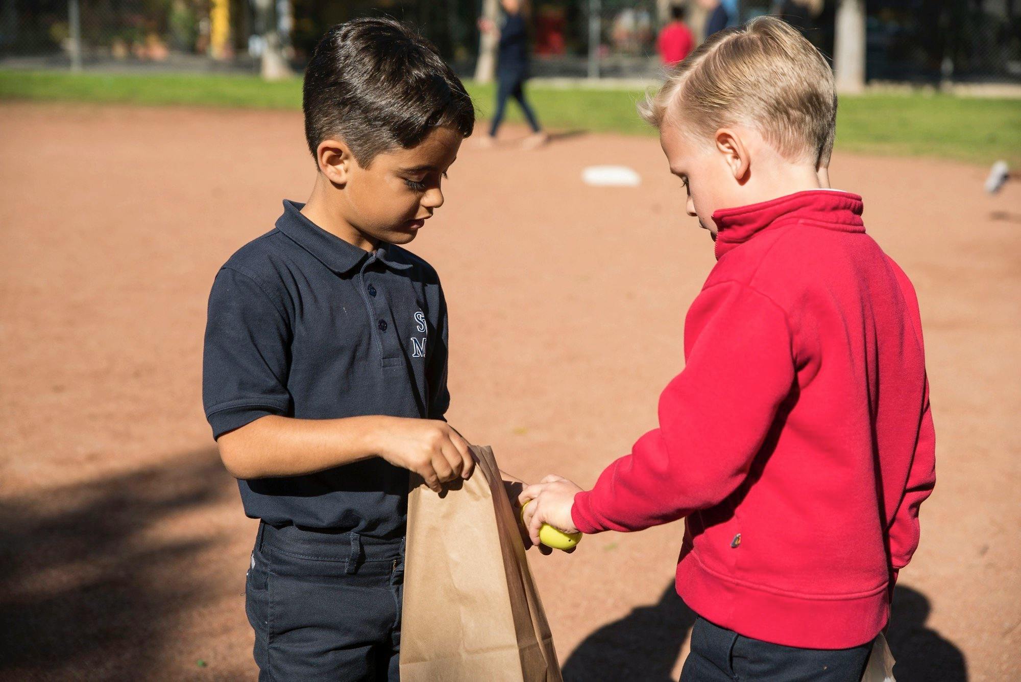 Child in blue shirt holding a paper bag, child in red shirt puts an apple in it