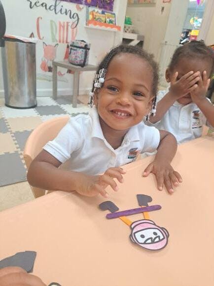 Child smiling at a table with another child covering their face