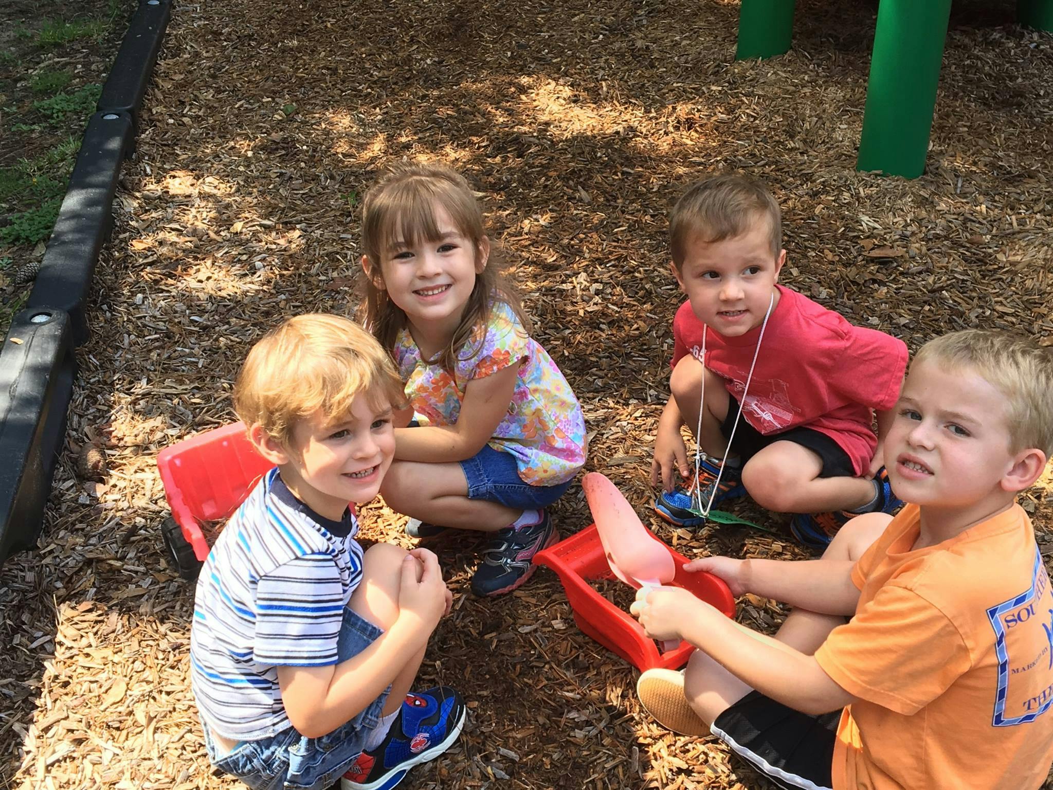 Four children playing on wood chips with red toy shovels