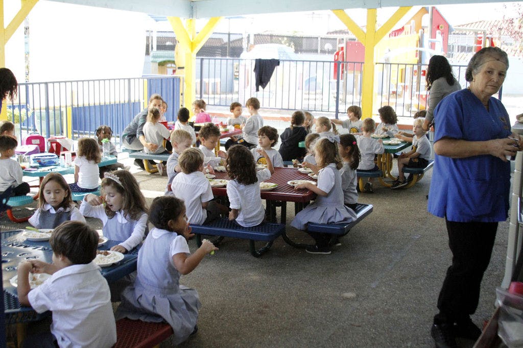 Many children eating at tables with an adult standing nearby