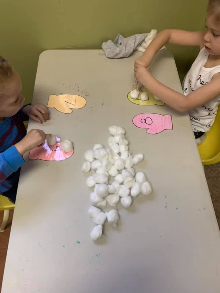 Two children doing a craft with cotton balls and paper cutouts at a table
