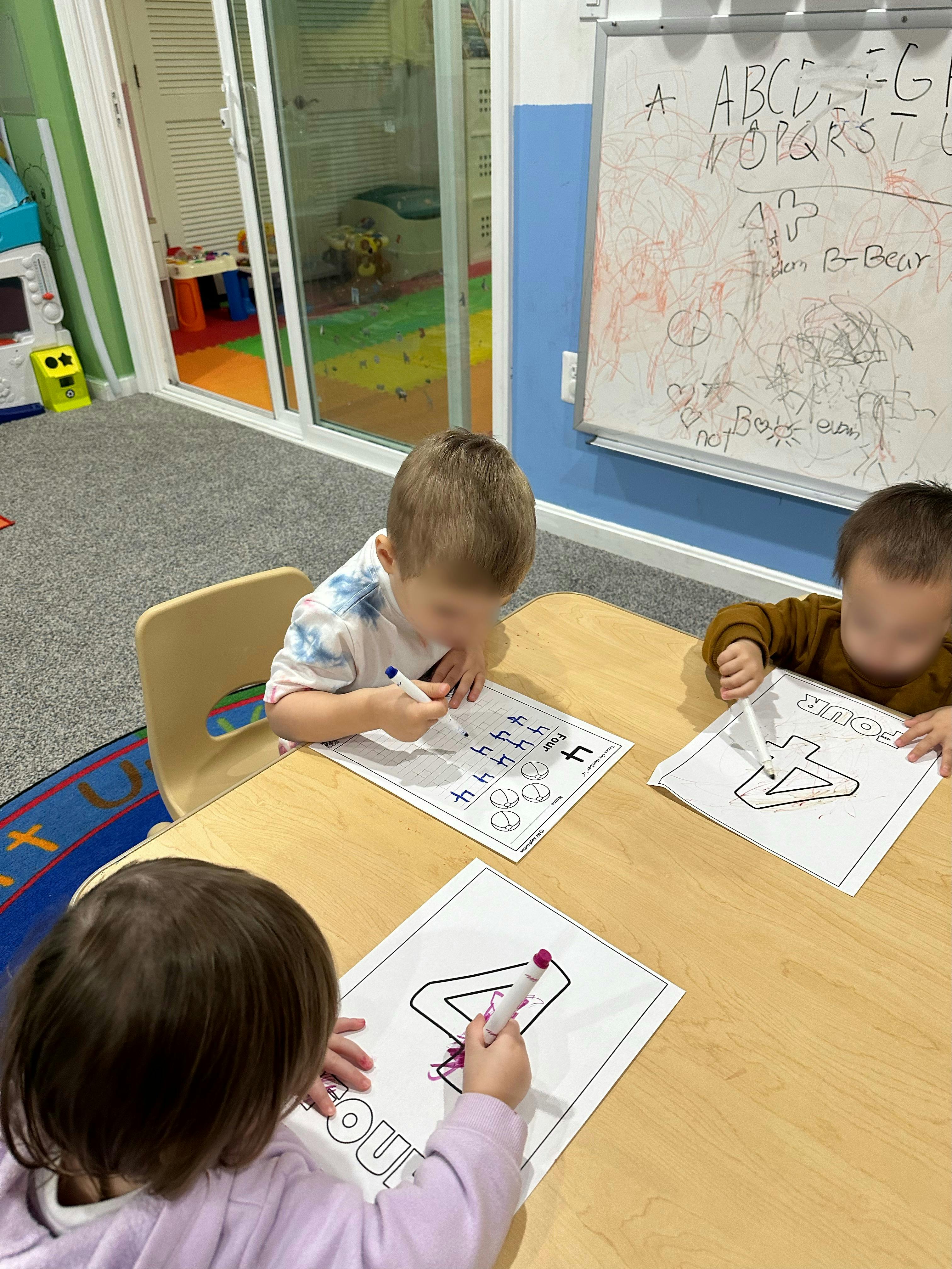 Three children coloring number four worksheets at a table in a classroom