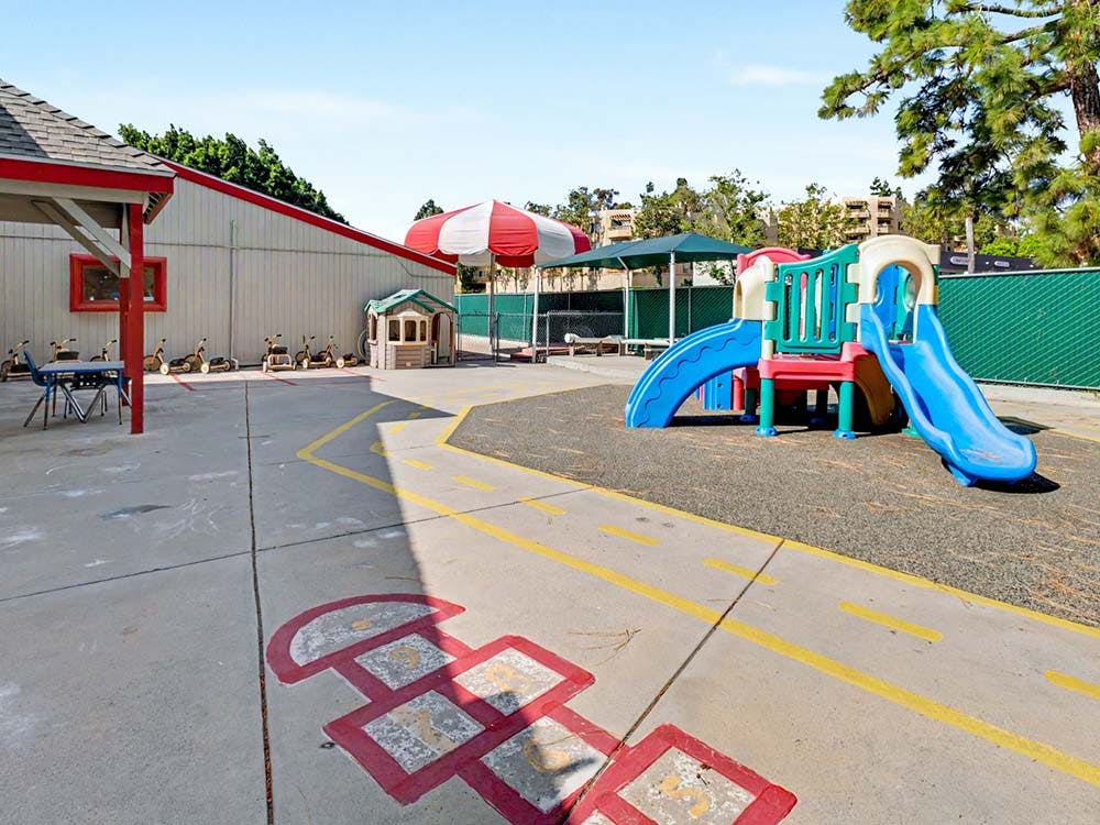 Playground with slides, building, umbrella, and painted hopscotch on pavement