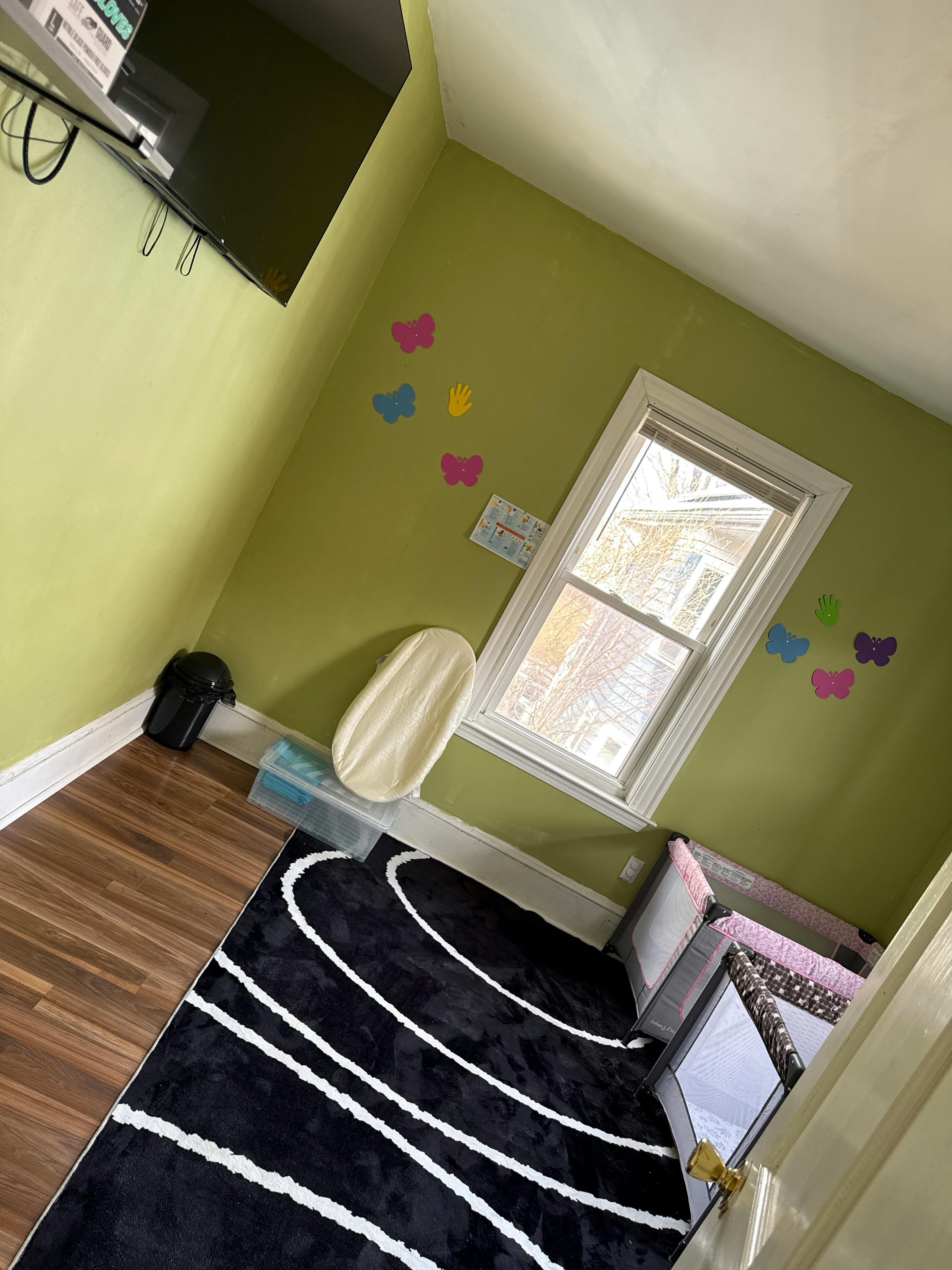 A room with light green walls, a window, a pack-and-play, and a black rug