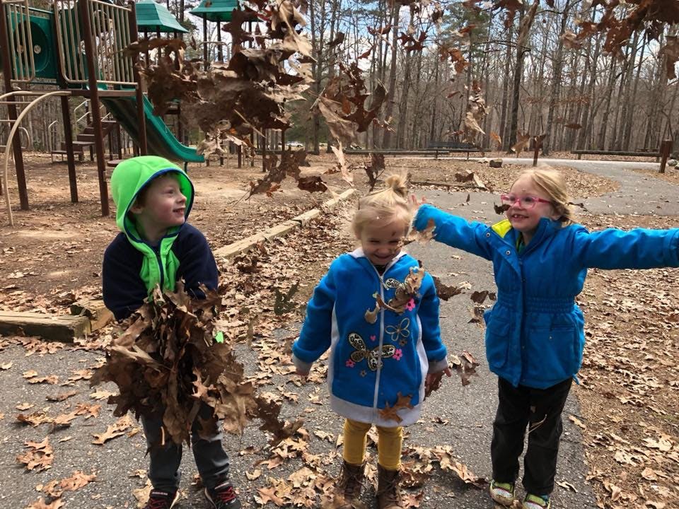 Three children playing with fallen leaves outdoors near a playground