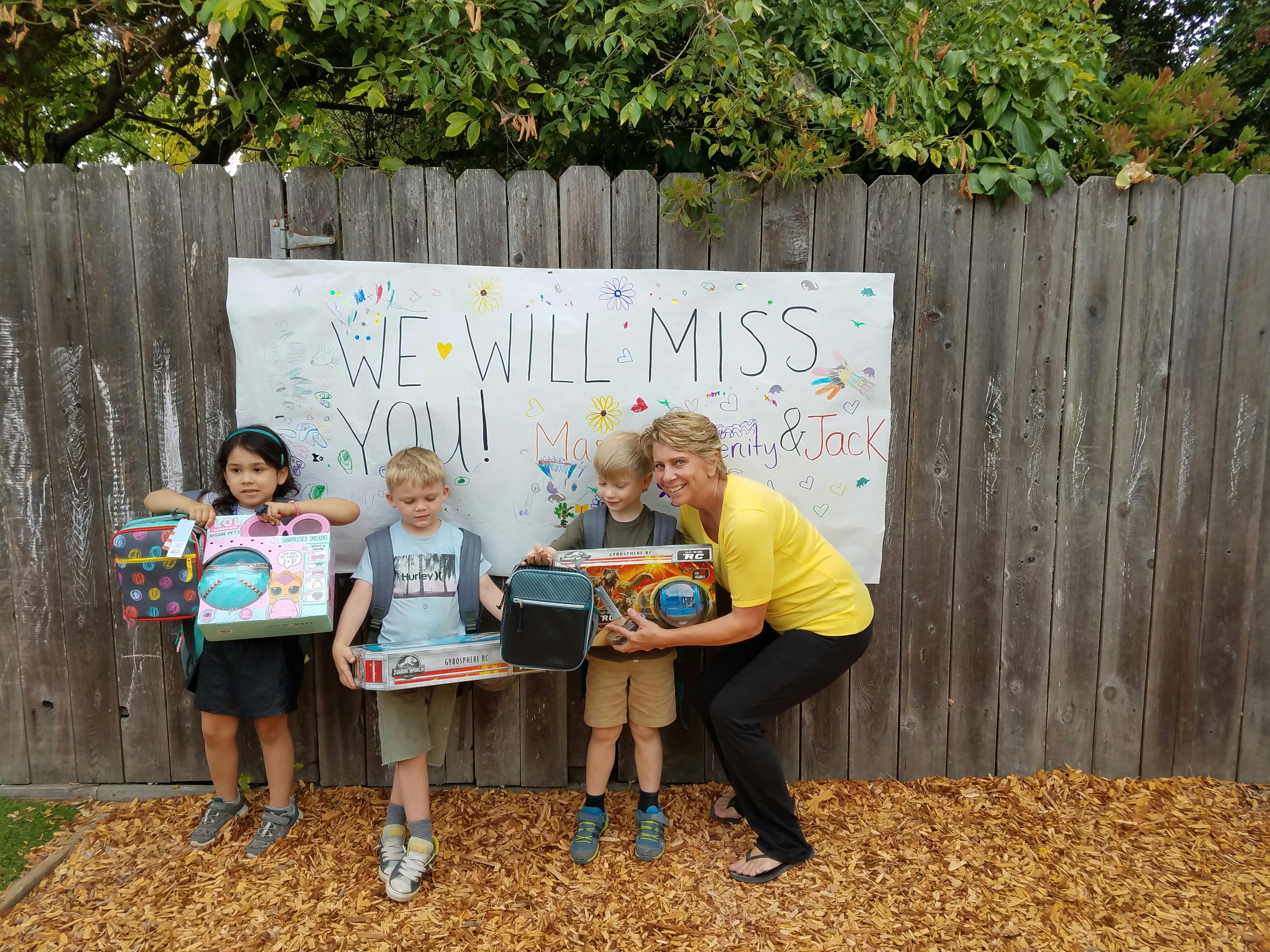 Adult and three children holding gifts in front of a "We Will Miss You" sign