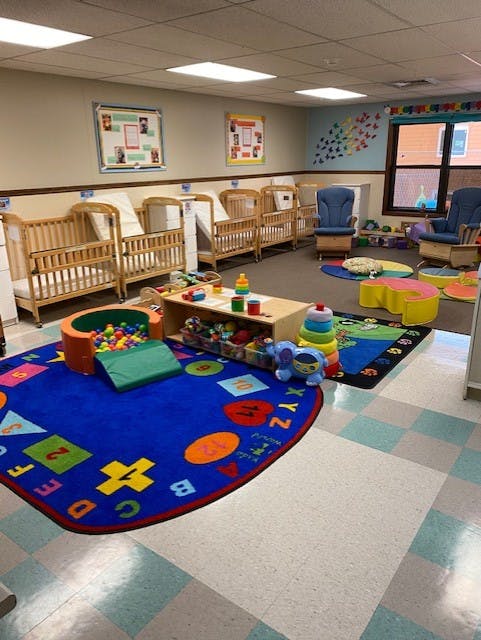 daycare classroom with multiple cribs, blue chairs, and a play rug