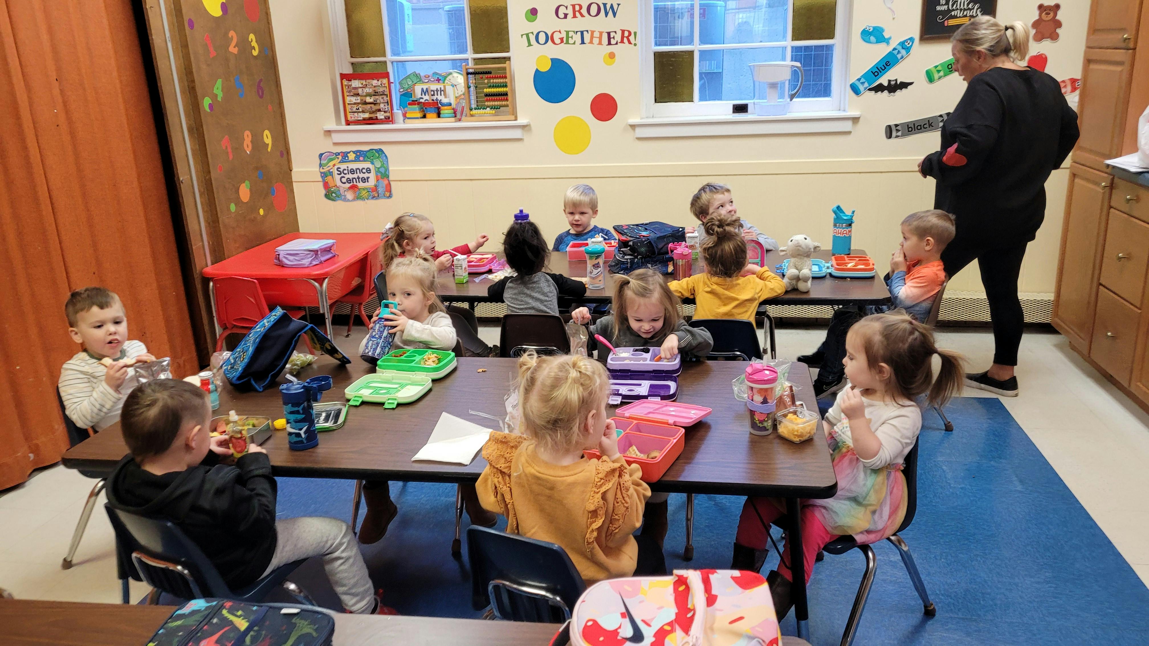 Children sitting at tables in a classroom with an adult standing nearby
