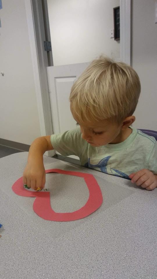 Child working on a craft with a red heart shape on a table