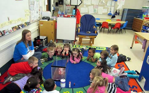 Adult and children sitting on a rug in a classroom looking at a screen