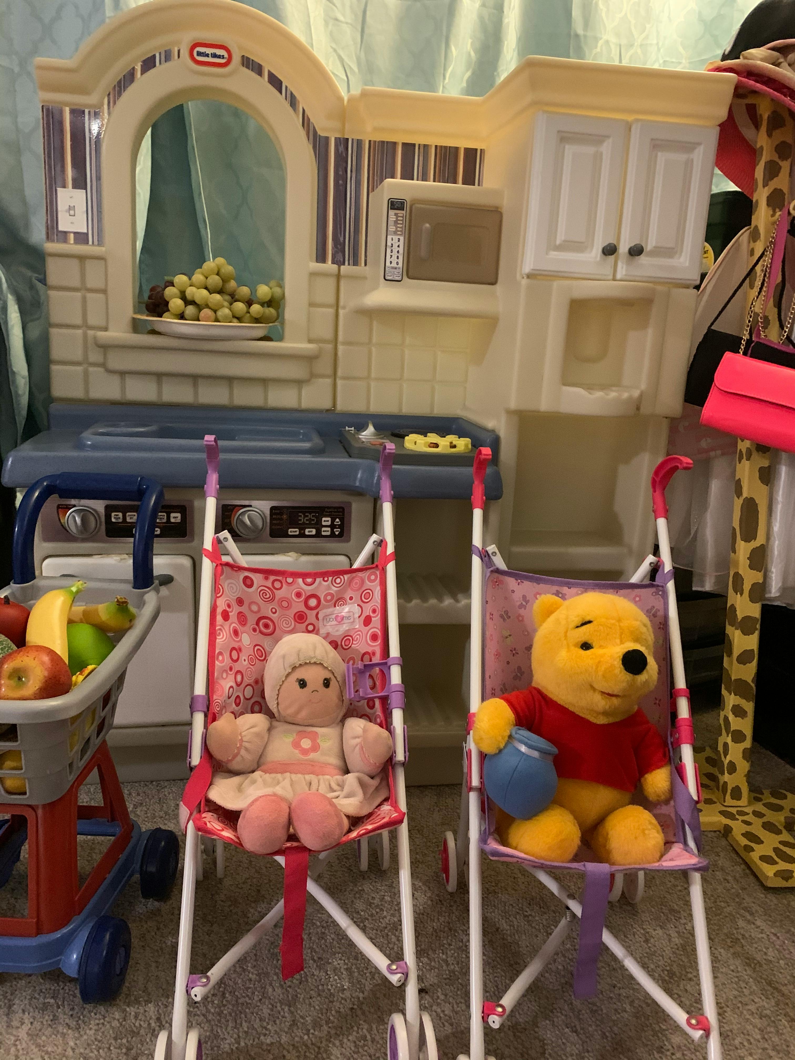 Dolls in strollers in front of a toy kitchen with a shopping cart of fruit