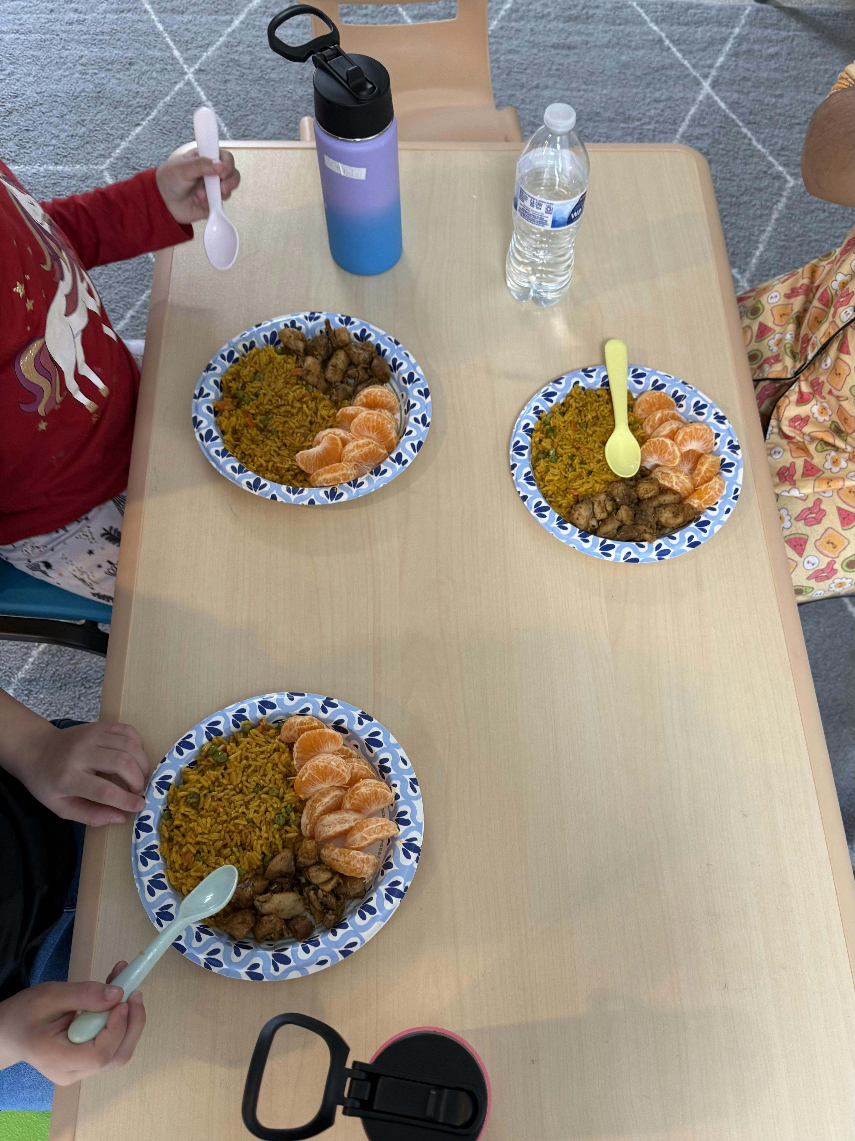 Children eating chicken, rice, and mandarin oranges on paper plates
