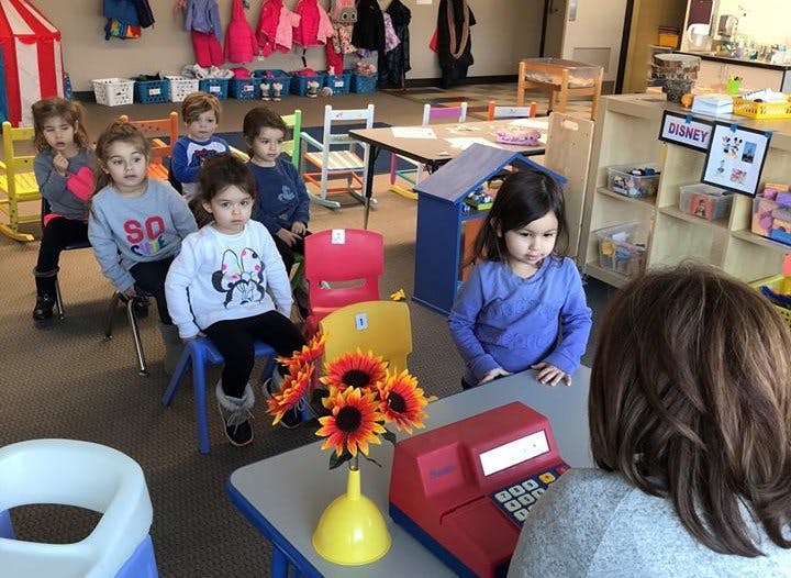 Children in a classroom, some sitting in chairs, one standing at a table