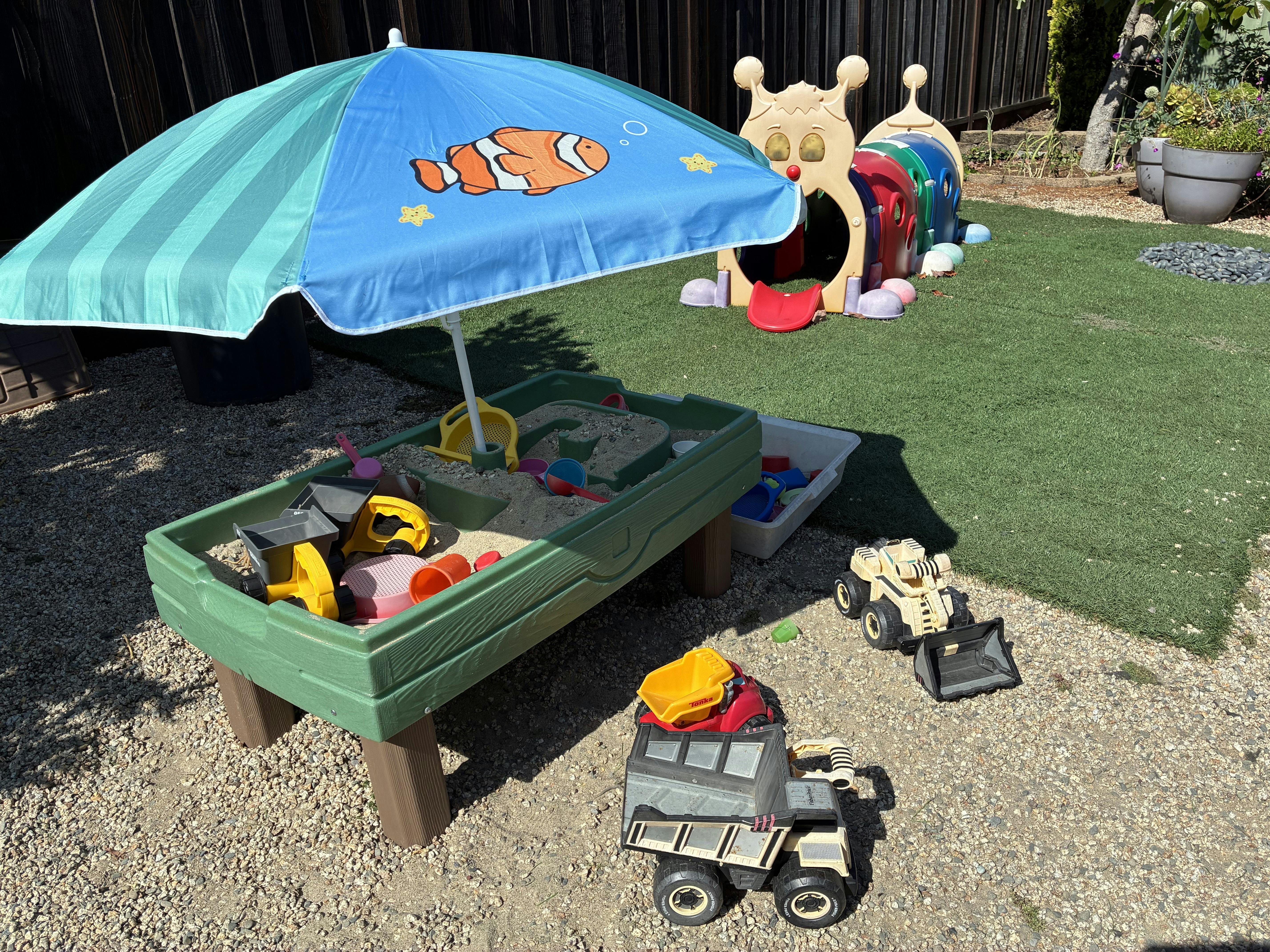 Sandbox with toys under an umbrella, caterpillar tunnel, and toy trucks