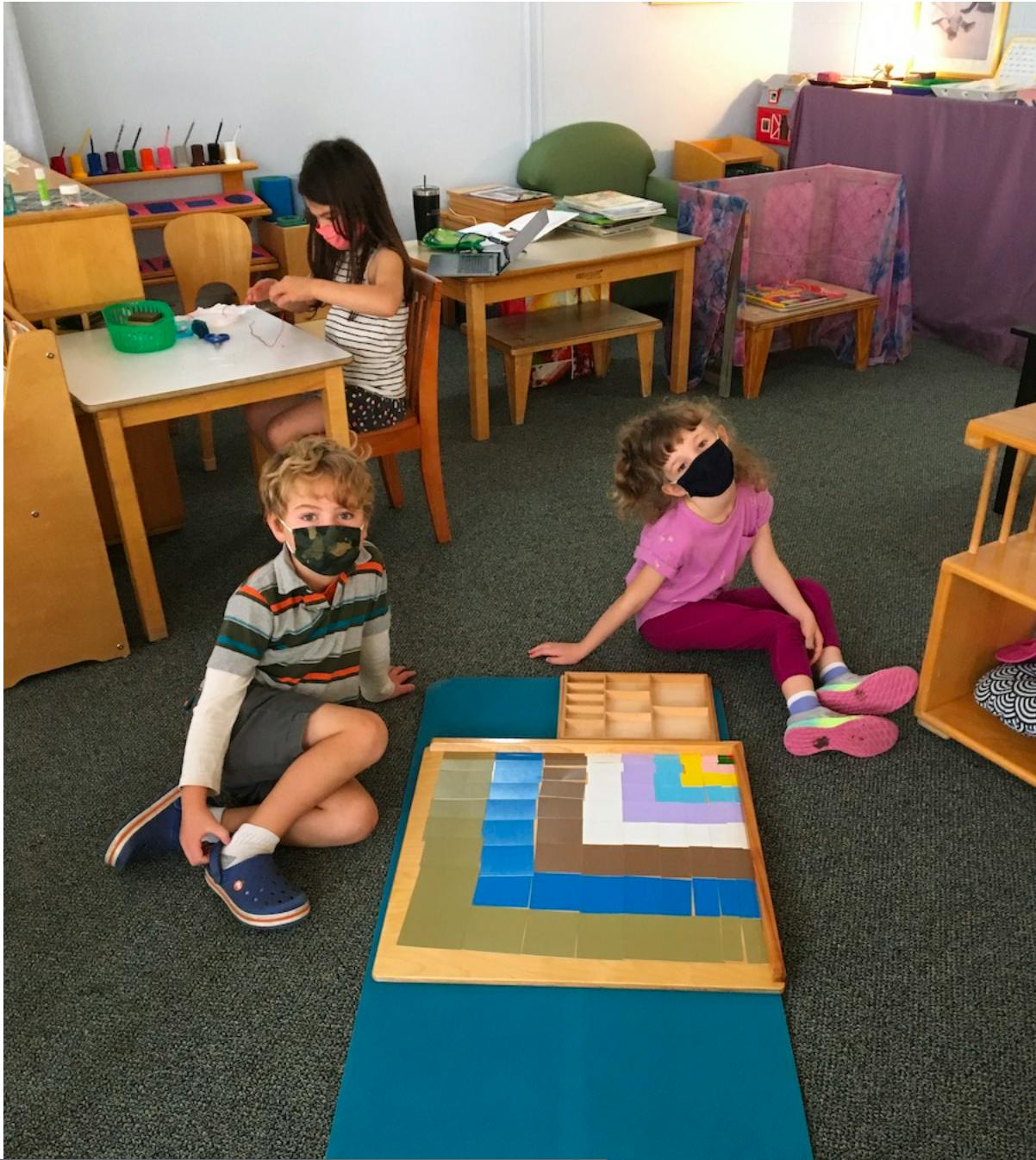 Three children in face masks playing with learning materials in a classroom