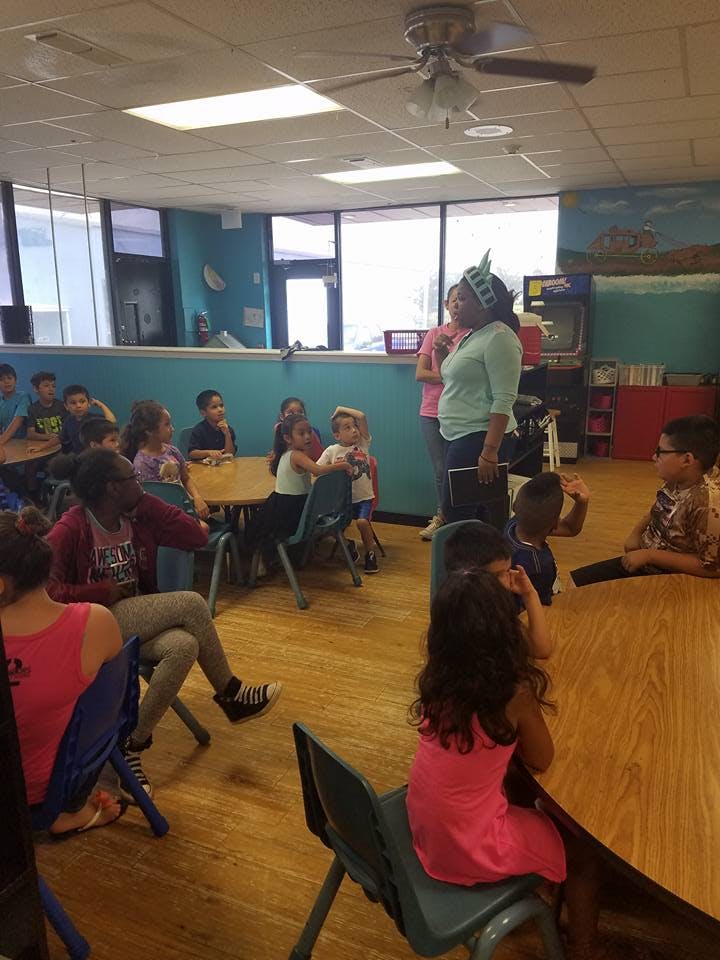 Adult speaking to a group of children in a classroom setting with tables and chairs