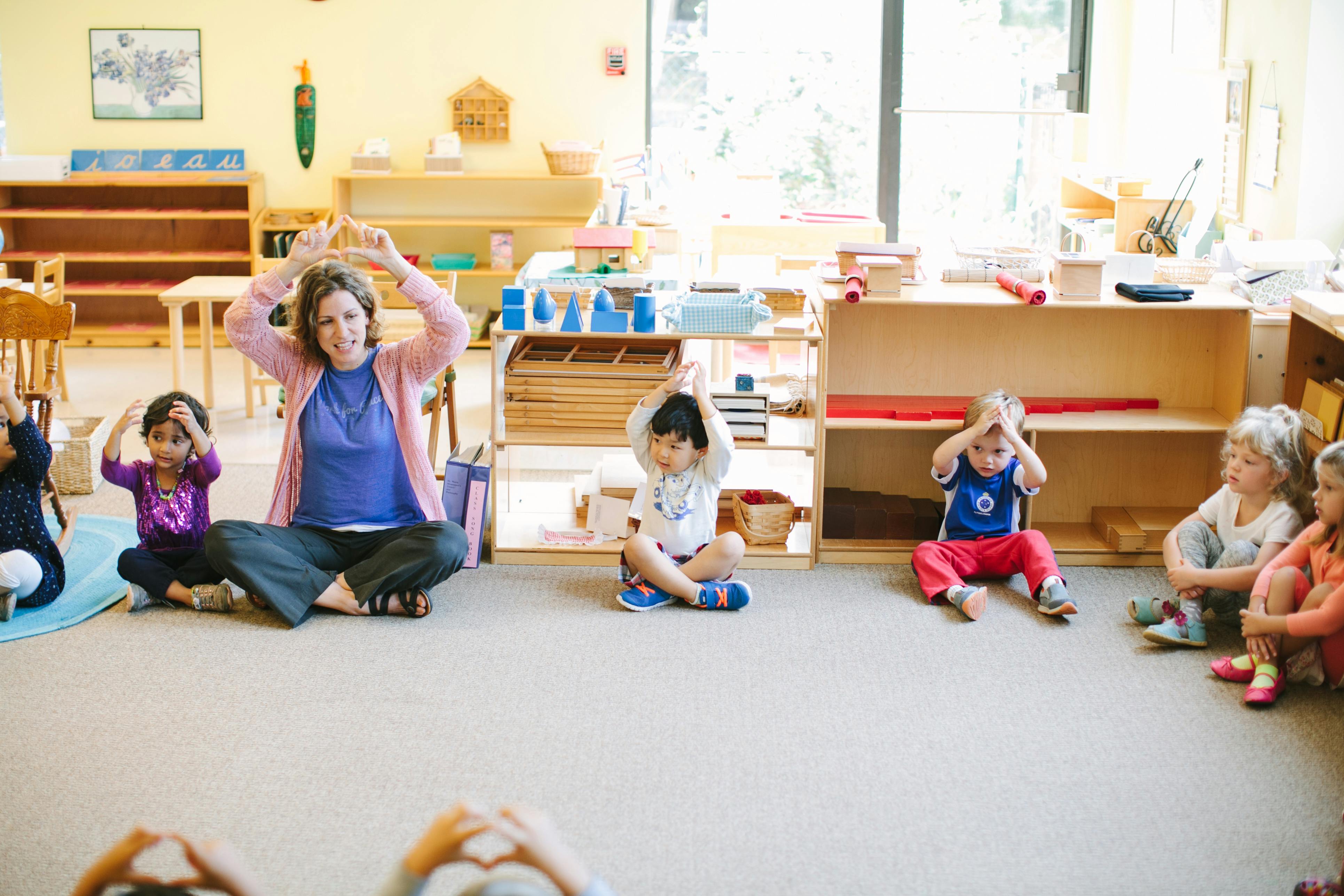 Adult and children sitting on floor with hands raised in a classroom