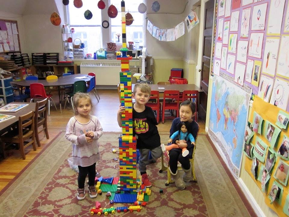 Three children standing around a tall structure made of colorful building blocks in a classroom