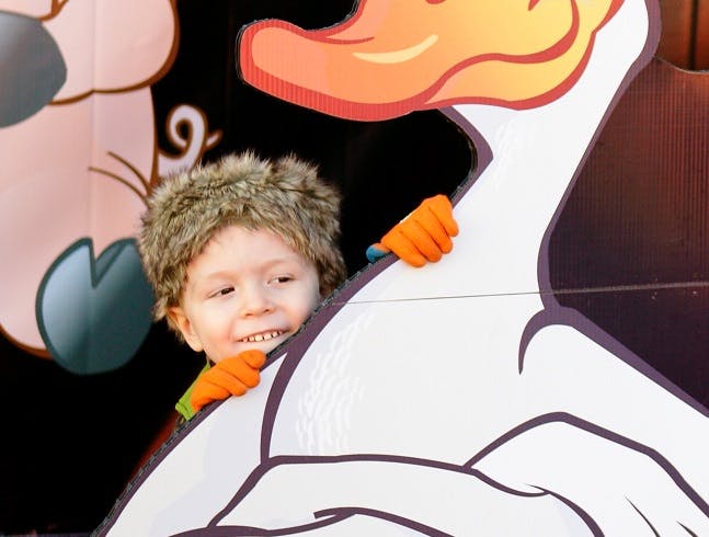 Child in a furry hat peeking through a cutout of a cartoon goose